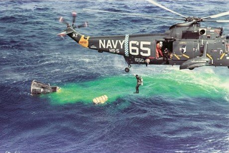 Image of a diver jumping into the ocean out of a helicopter that's hovering over a spacecraft floating in the water with a large area of green dye.