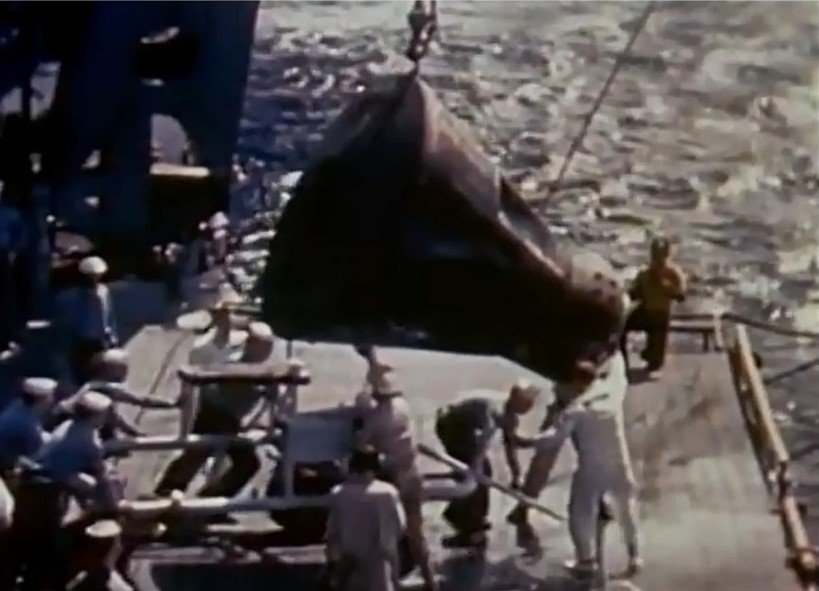 Sailors maneuver a cone shaped capsule hanging from a crane onto the deck of a ship.