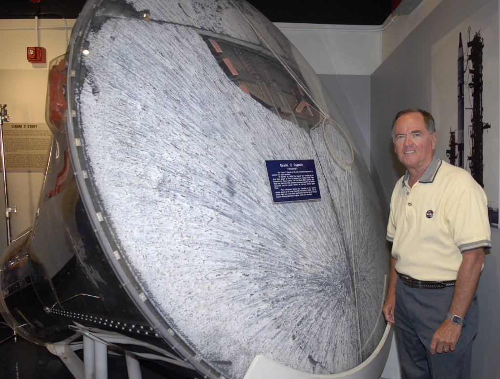 A man wearing a yellow shirt stands next to the hear shield end of a capsule on display in a museum.