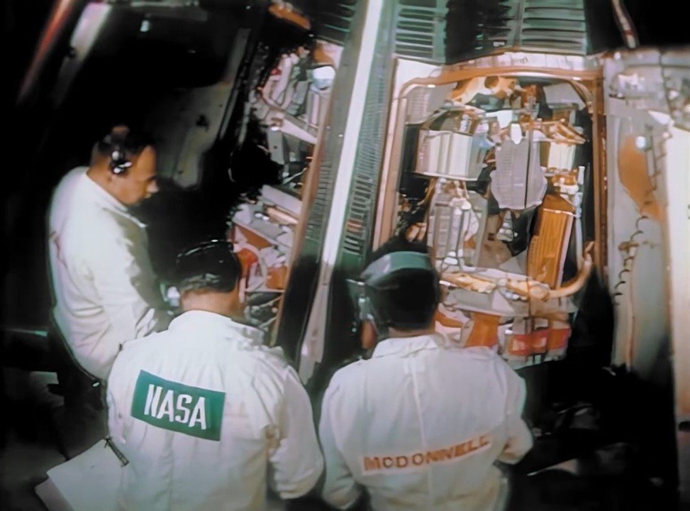 White coated technicians working around a spacecraft with two hatches open and instrumentation can be seen inside the spacecraft.