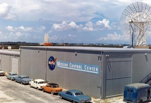 Image of a low building with the NASA meatball logo and the words "Mission Control Center" written in big letters, and several cars parked outside the building.