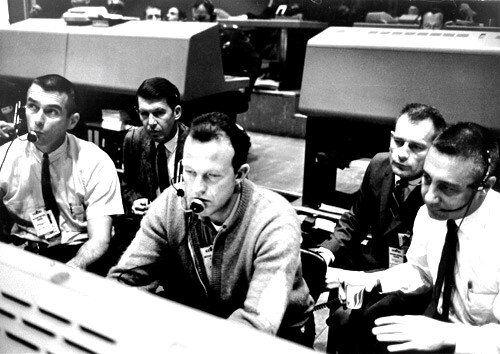 A group of five men in white shirts and ties hunched over a console in a control center.