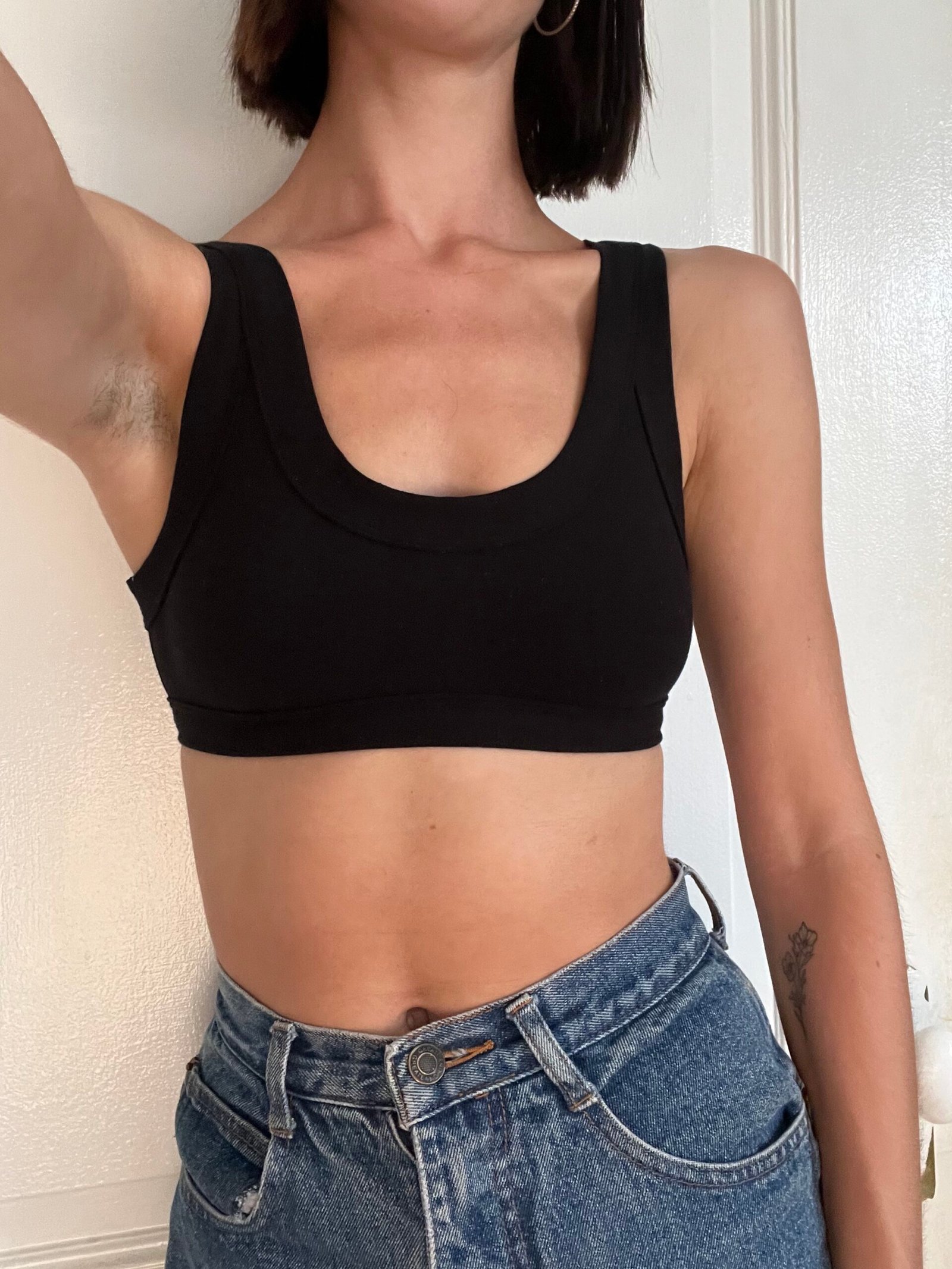 Person wearing a black crop top and high-waisted blue jeans, taking a selfie in front of a white wall.