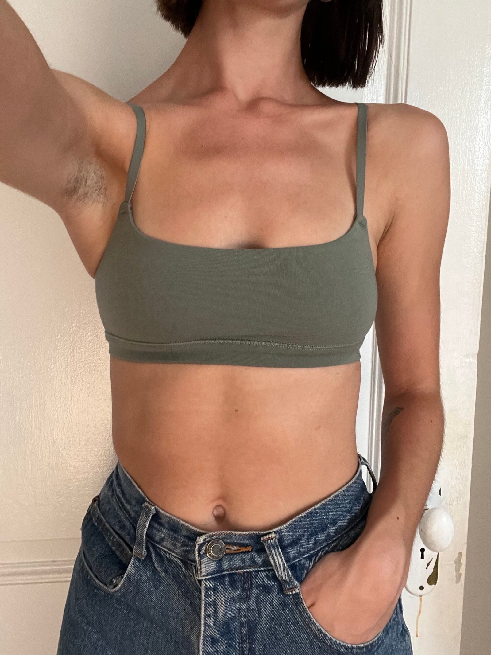 Person wearing a green bra top and blue jeans is taking a selfie. Their left arm is raised, and they have a visible tattoo on their forearm.