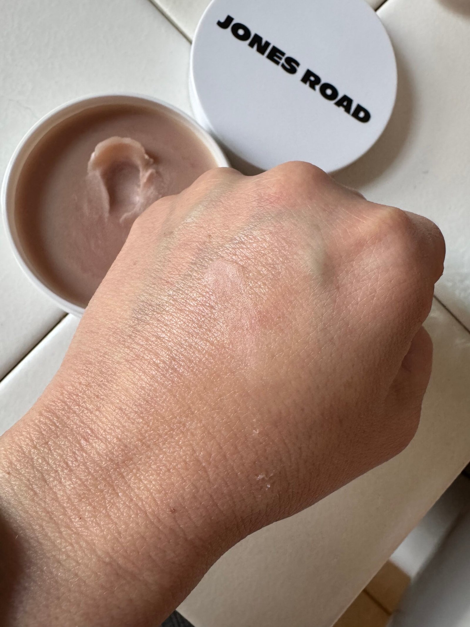 Close-up of a hand with a light makeup application, showcasing the natural finish alongside an open container of Jones Road product on a tiled surface