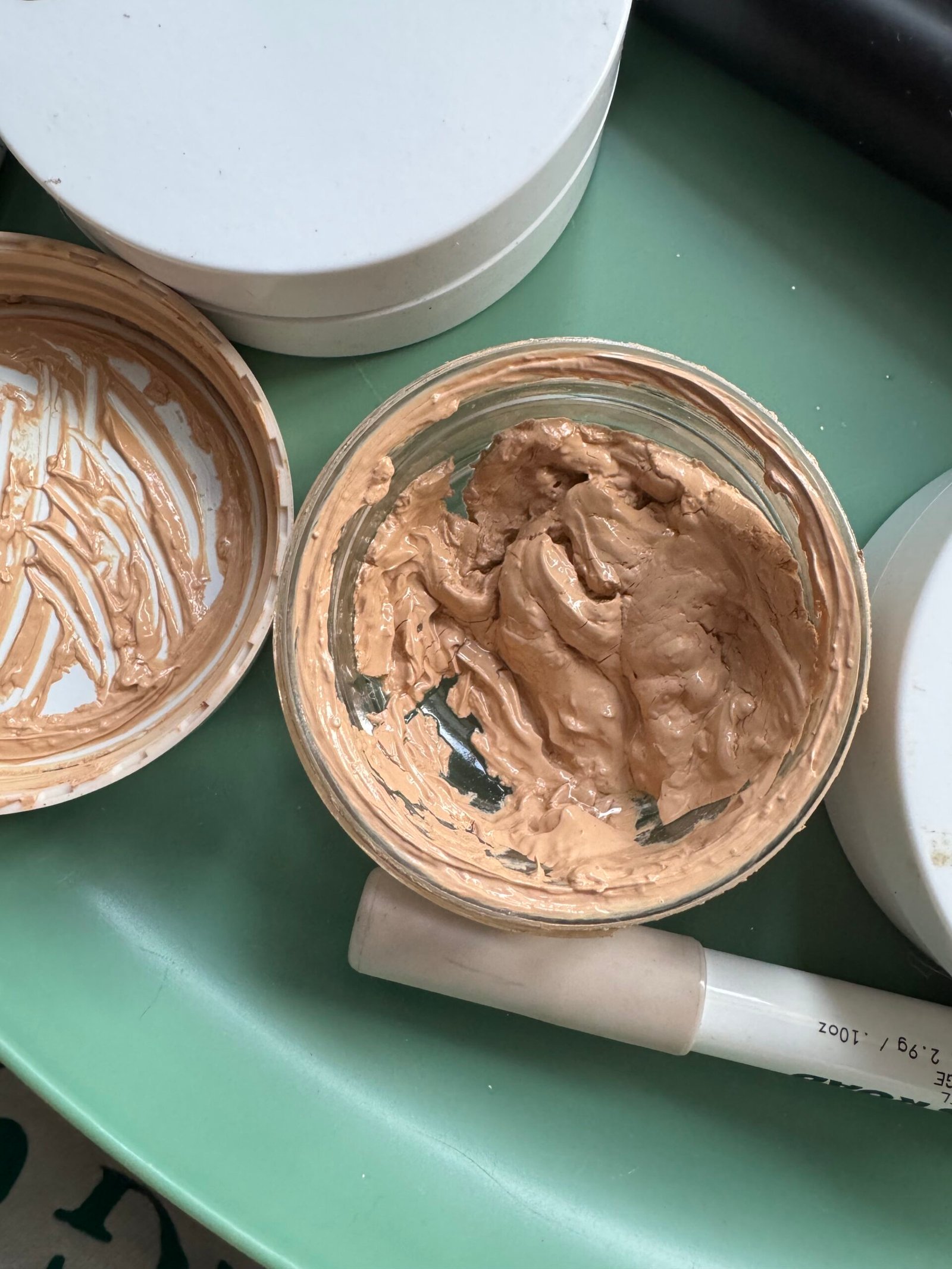 An open jar of What the Foundation from Jones Road sits on a fresh green surface, with some product used and its white lid resting beside it.