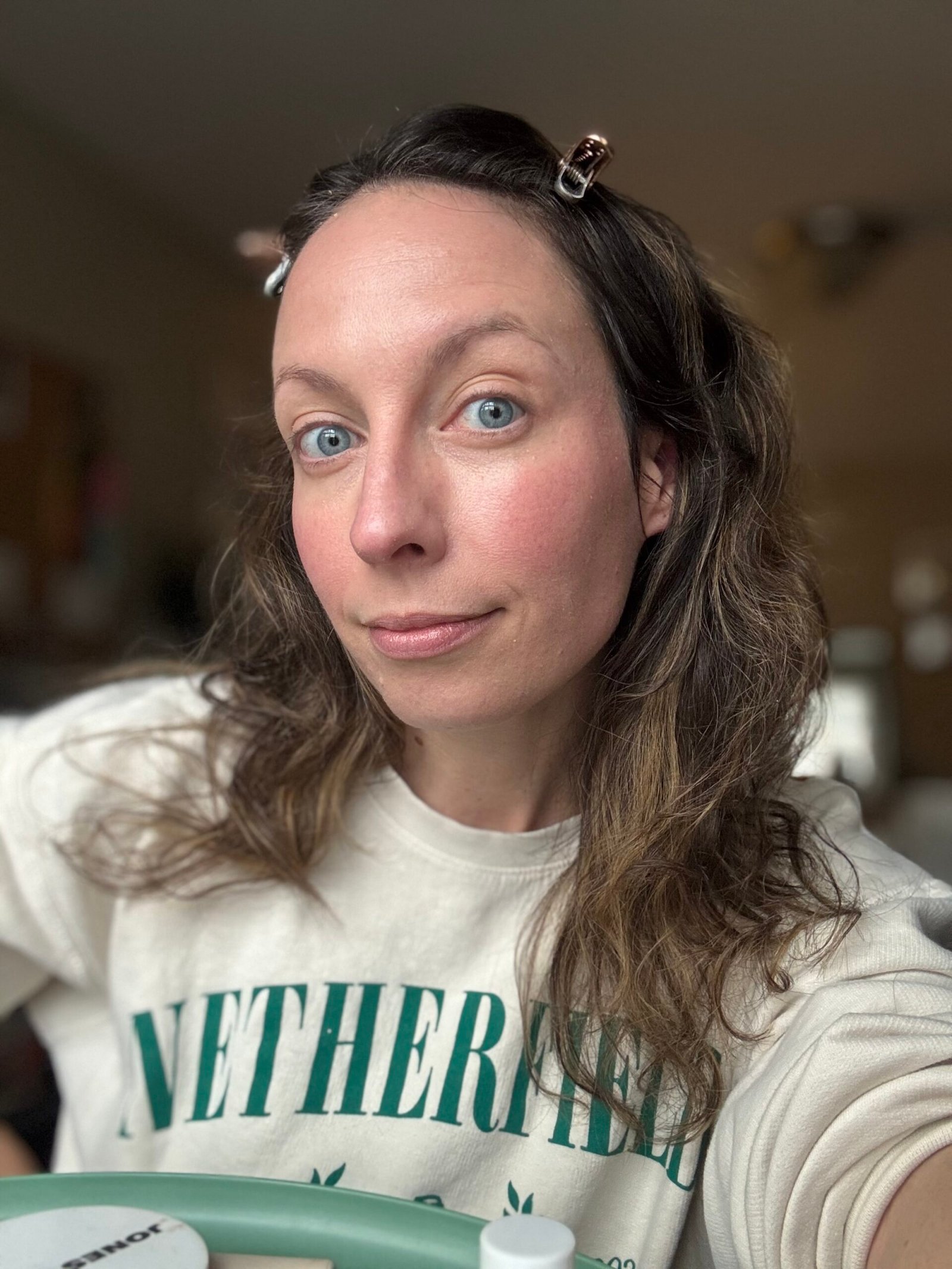 A person with long hair clipped back is wearing a light-colored sweatshirt featuring green text, reminiscent of Ilia's vibrant style.