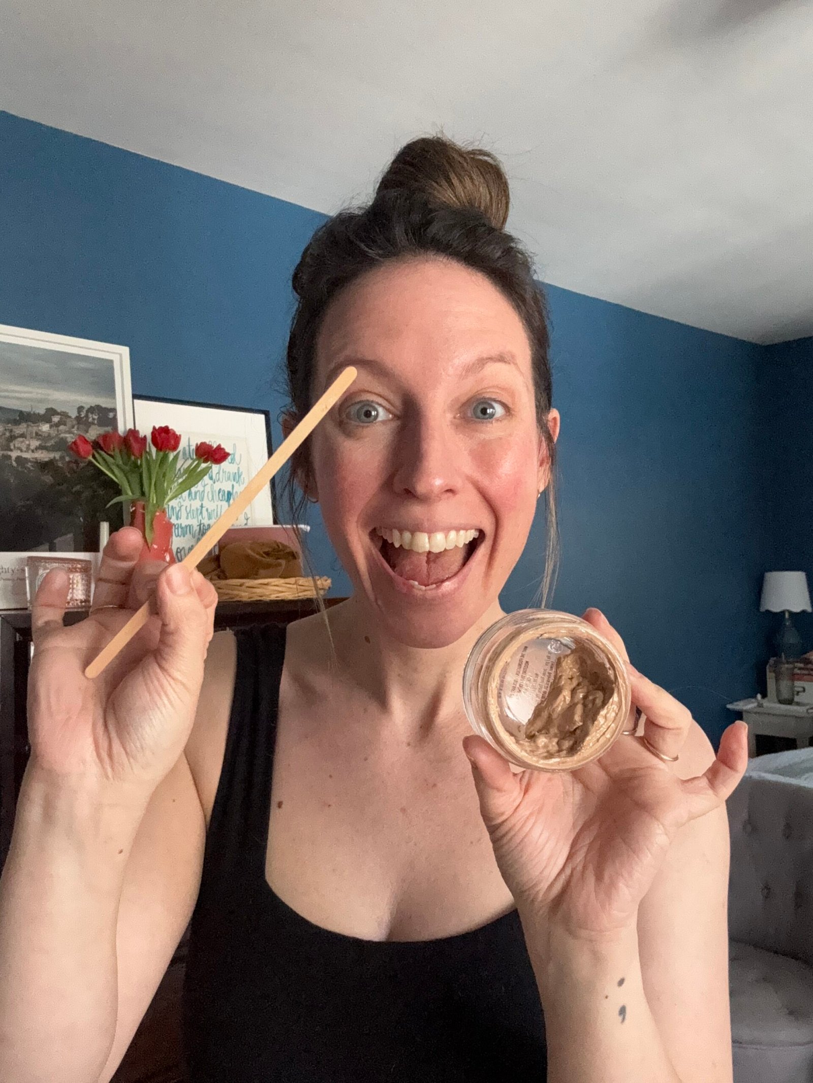 A woman in a black tank top smiles warmly, holding an empty container in one hand and a wooden stick in the other. 