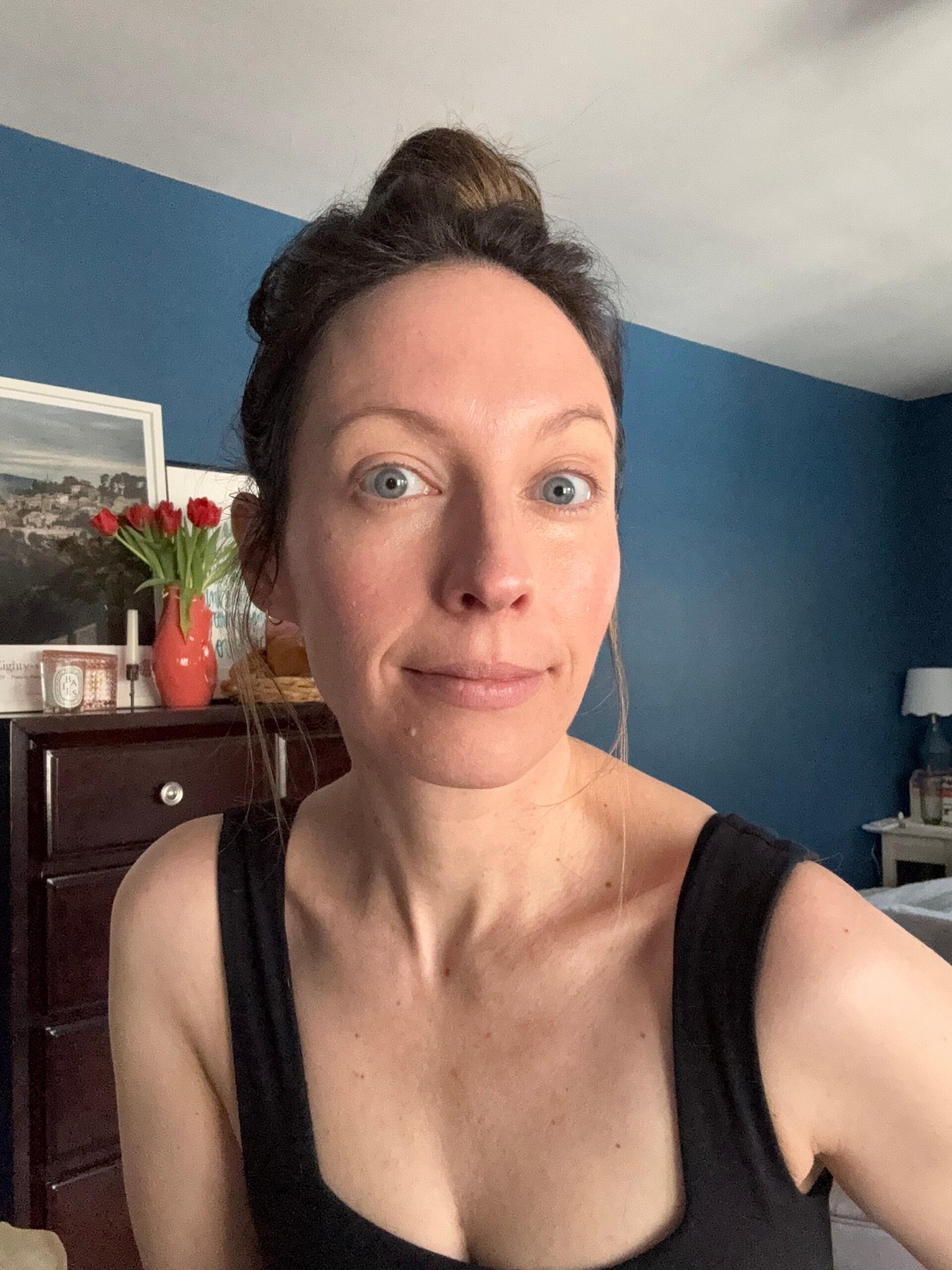 A person with light eyes and a topknot hairstyle, resembling an Ilia character, is wearing a black top while sitting indoors in front of a dresser adorned with a vase of vibrant red flowers.