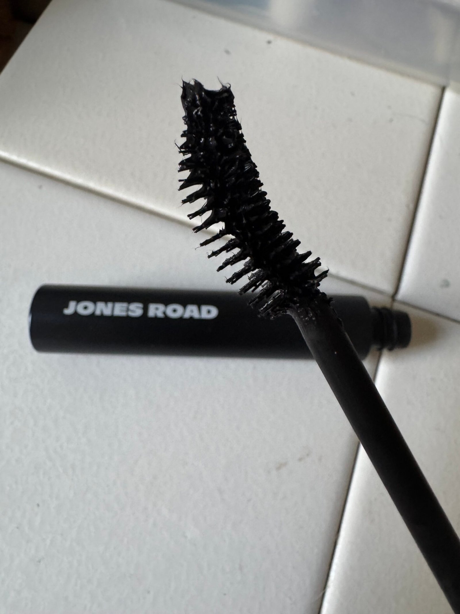 A black mascara wand with a curved brush is in the foreground, capturing attention. The sleek black tube with "Jones Road" elegantly written on it sits stylishly on a white tile surface.