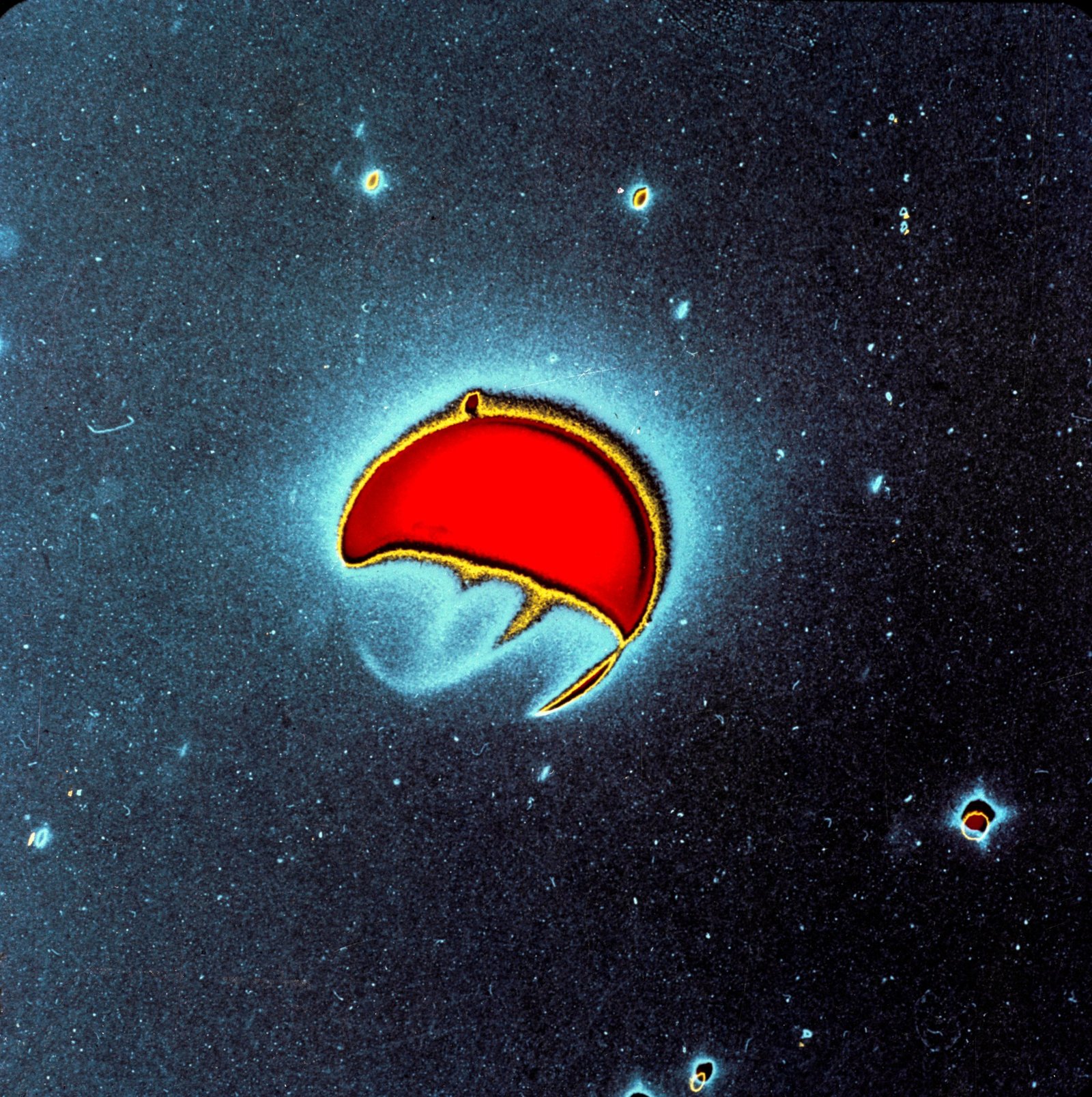 In this enhanced-color photo, Earth is represented in shades of aqua blue, red, and yellow. Earth's atmosphere is a bright blue haze, while the planet itself is red with a yellow outline on the sunlit side. Nearby stars are visible as blobs with similar colors to Earth.