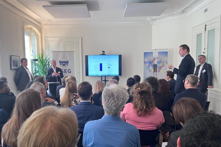 Room full of seated people watching a presentation on a screen at the front of a white room.