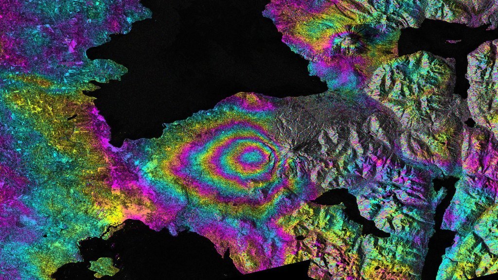 A satellite image of the Calbuco volcano in Chile with a swirling pattern of vibrant colors highlighting the land deformation caused by its 2015 eruption. The image showcases the impact of the volcanic activity on the surrounding landscape.