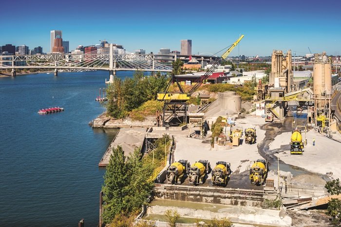 A cement factory stands beside a river in Portland, Oregon, near a suspension bridge.