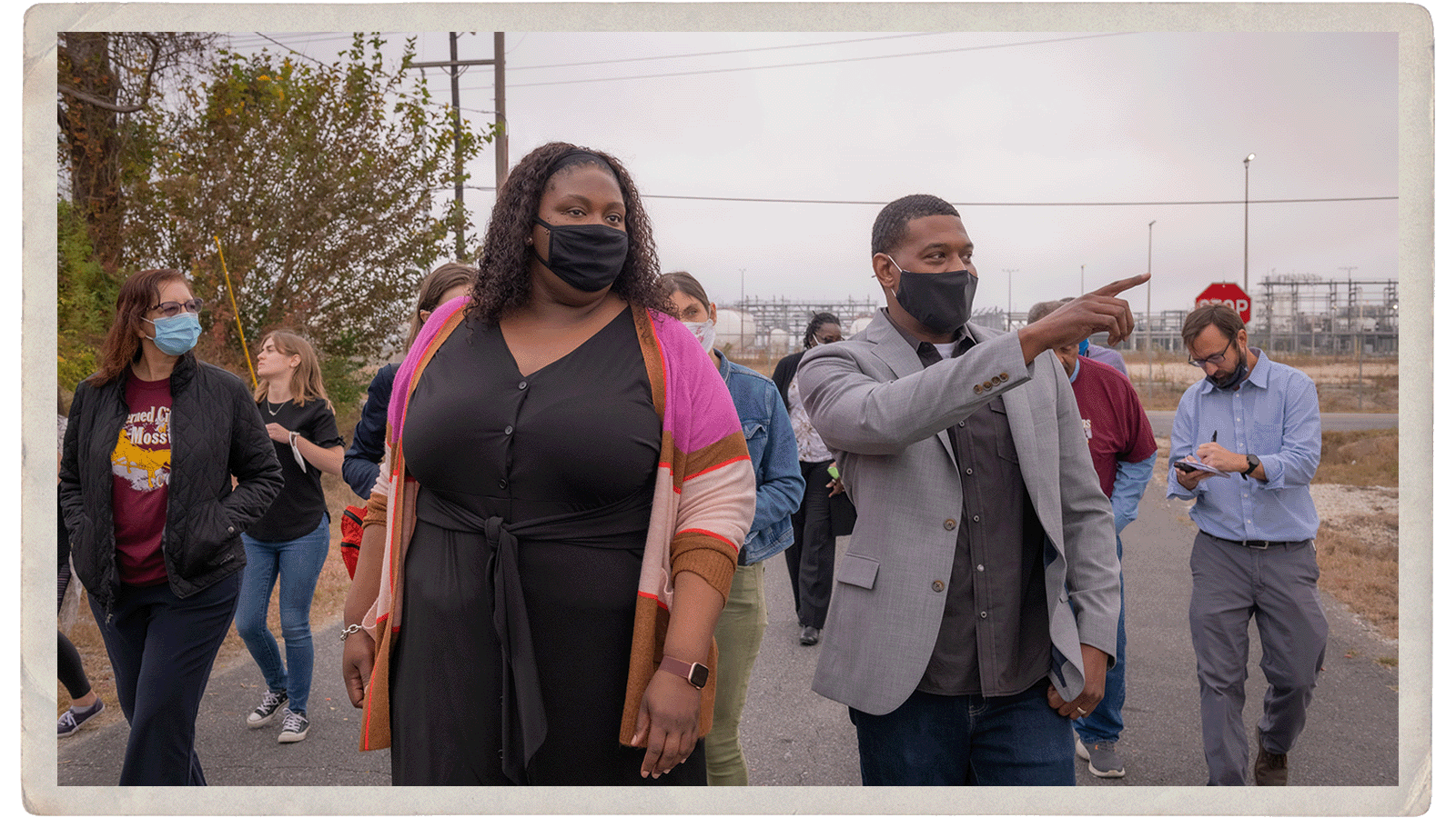 A group of people walk along a tree-lined street. One, Michael Regan, is pointing at something off-scene.