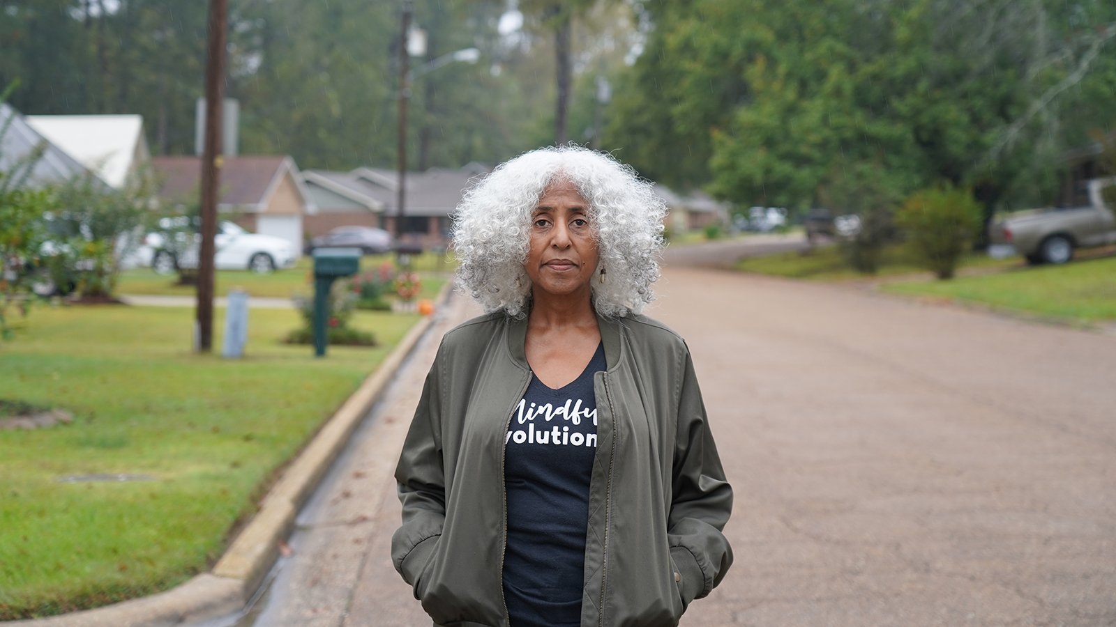 A woman with gray hair looks directly into the camera while standing in front of a residential street