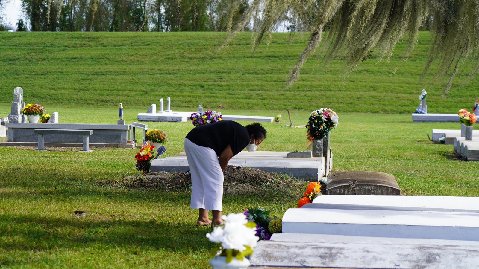 A woman bends over a grave marker with flowers in a cemetery lawn