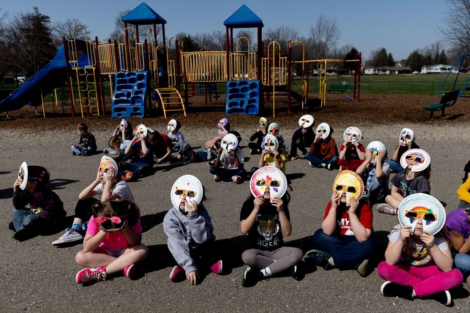 Children sitting on the asphalt at a playground and looking up at the sky. Each one was wearing special solar viewing glasses that were attached to a decorated paper plate.