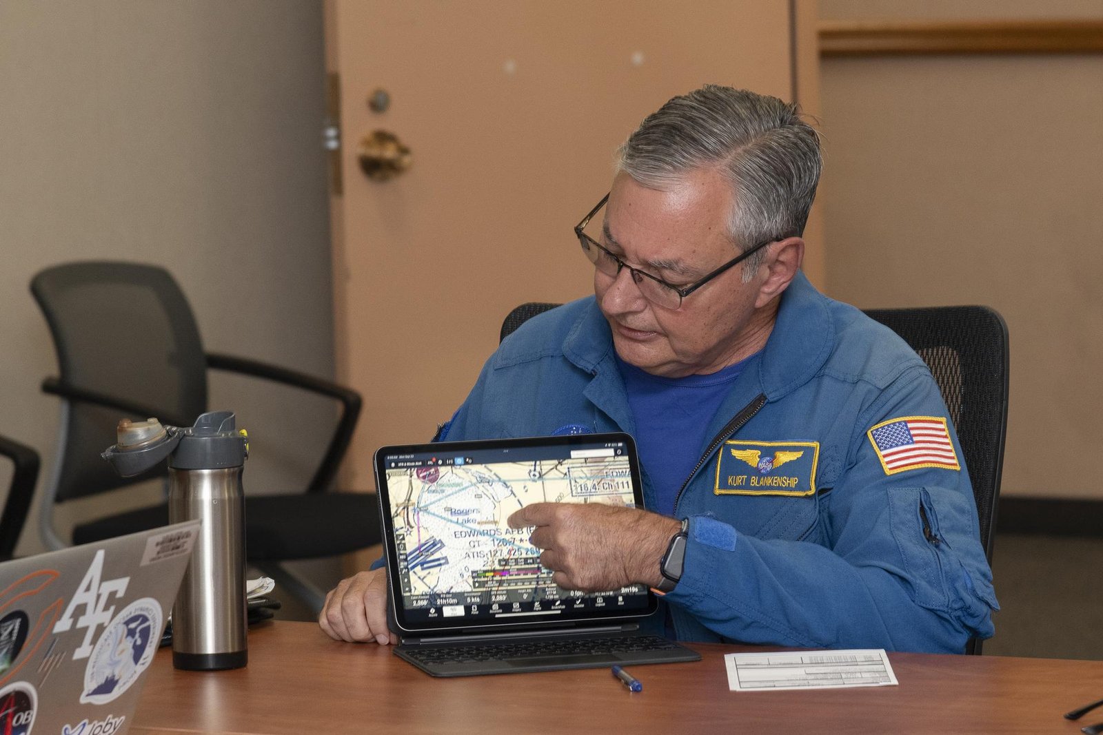 In a briefing room at NASA’s Armstrong Flight Research Center, in Edwards, California, NASA pilot Kurt Blankenship wears a blue flight-suit and sits at a brown desk to review flight plans on a rectangular flight tablet. The tablet displays a map of Edwards Air Force Base and Rogers Dry Lakebed with directional lines in light blue and flight zones designated in dashed lines and purple circles.