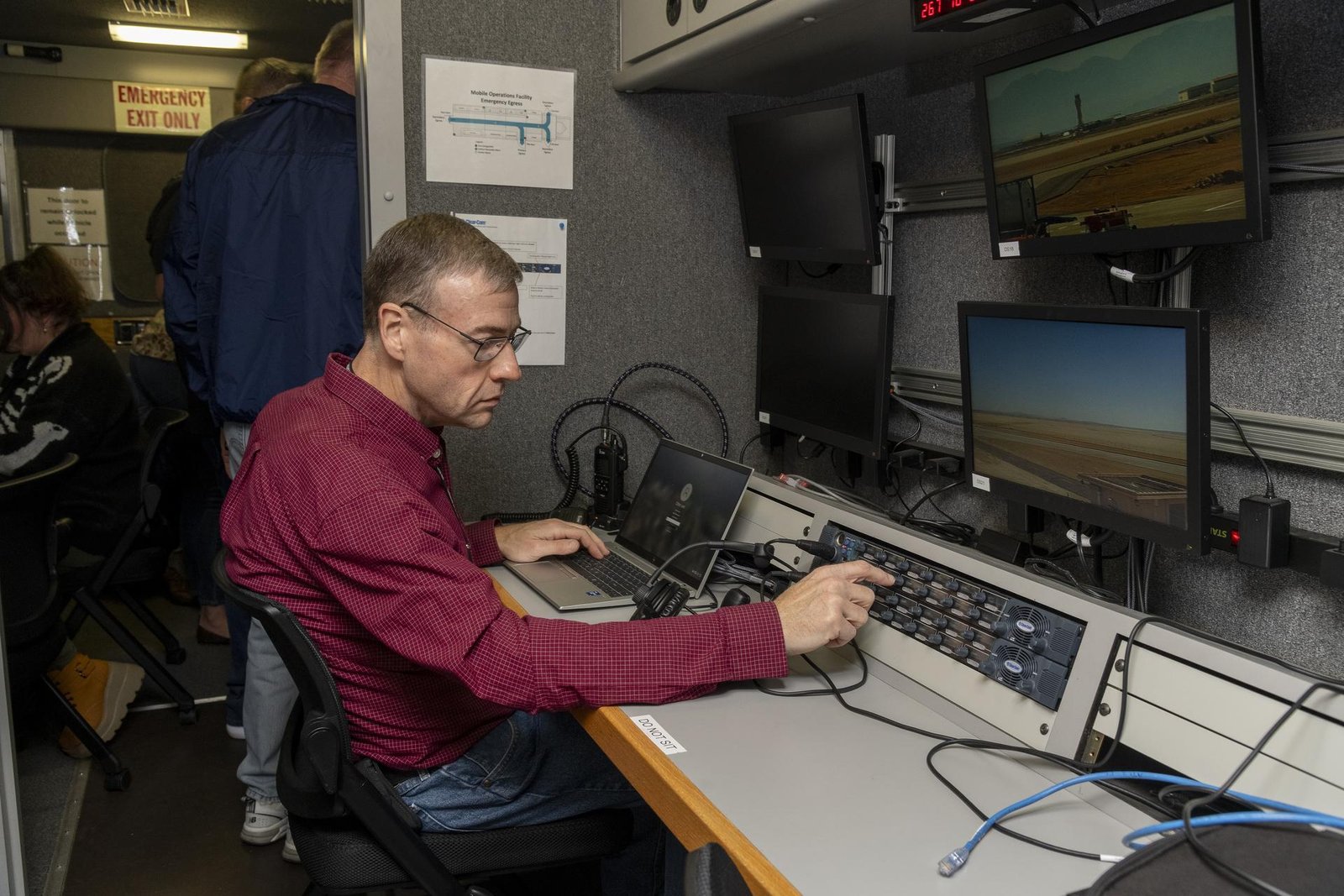 Wearing a dark red shirt, NASA researcher Dennis Iannicca, sits at a control monitor with three video screens, a laptop, and a control board with dials. The gray-colored control station is inside the Mobile Operations Facility, a large trailer that houses multiple computer workstations to monitor flight testing. The ADS-B research is being done at NASA’s Armstrong Flight Research Center in Edwards, California.