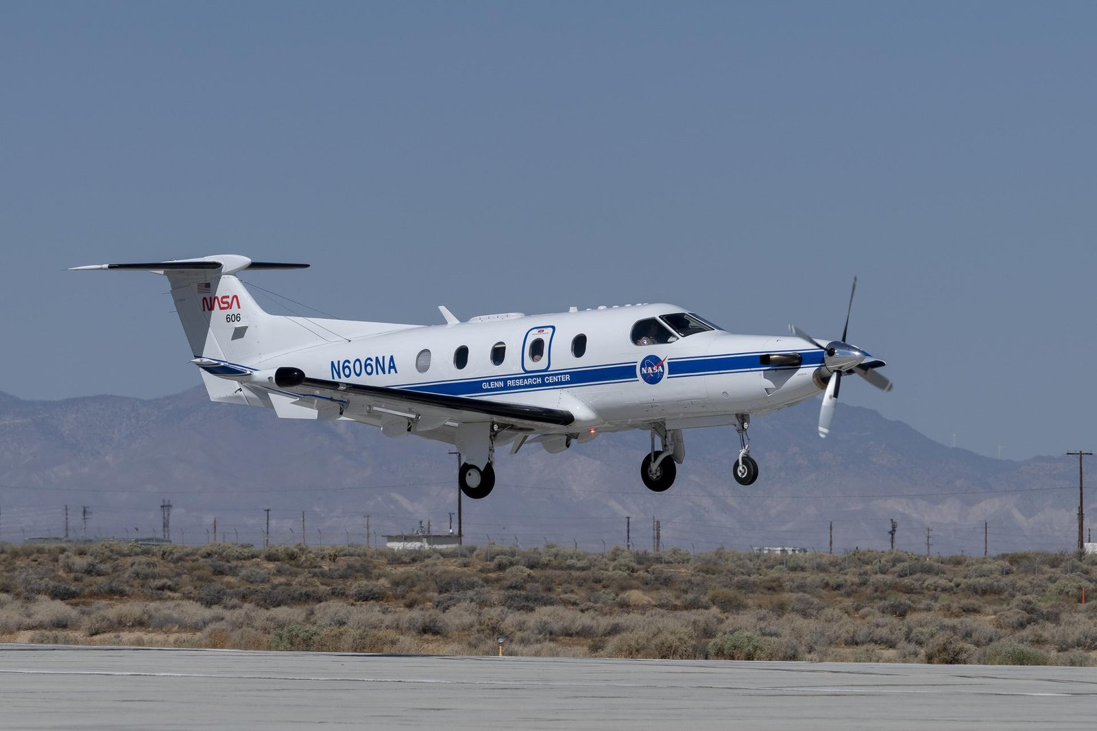 NASA’s Pilatus PC-12 flies over the runway at NASA’s Armstrong Flight Research Center. The white plane shines bright against the Mojave Desert landscape. The red NASA worm figures prominently on the tail of the plane and a blue stripe lining the fuselage with the NASA meatball logo under the pilot's window. The plane’s call sign, also in blue, N606NA is brightly painted above the blue stripe.