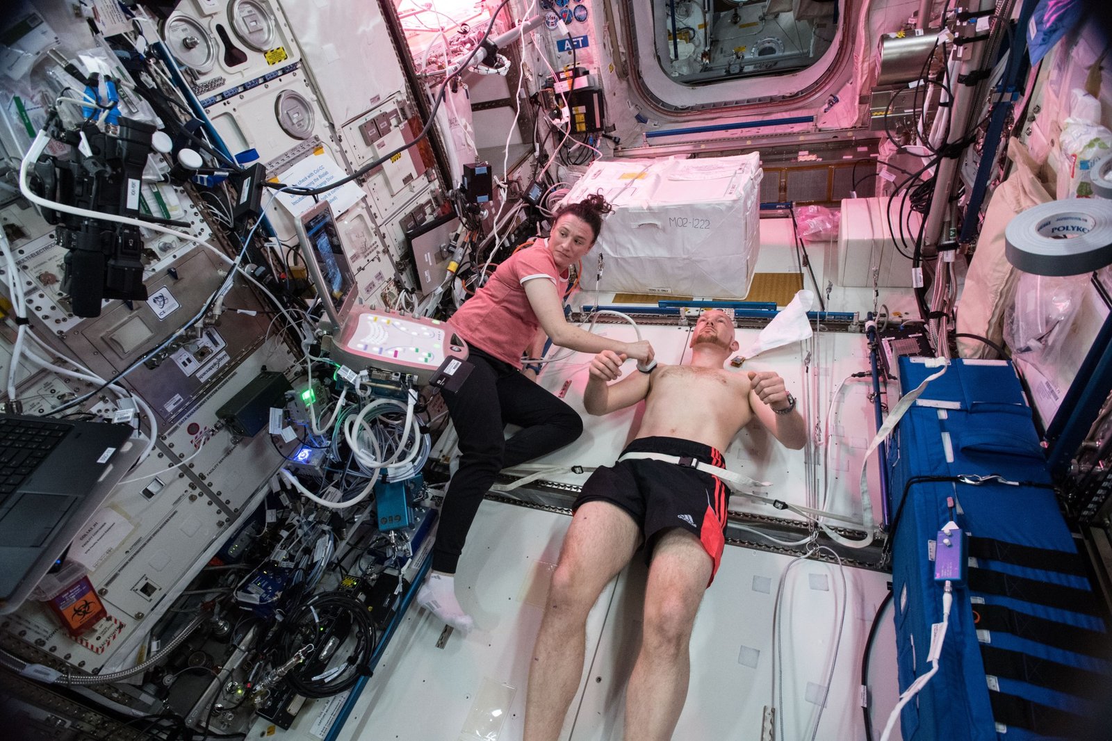 Gerst, wearing black shorts, lies on his back while Auñón-Chancellor holds a small white ultrasound device over his right upper arm. Auñón-Chancellor is wearing a pink shirt, black pants, and white socks. Her head is turned toward the screen of the ultrasound.