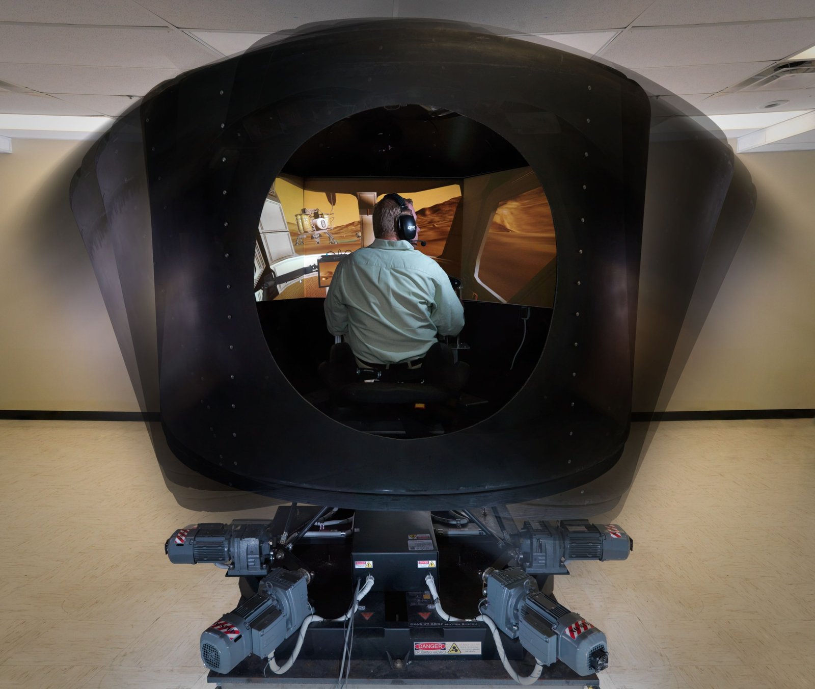 Dean, wearing a green shirt and a headset, is seated inside the cockpit-shaped black simulator, with his back toward the camera. Images of the surface of Mars are projected on screens in front of him.