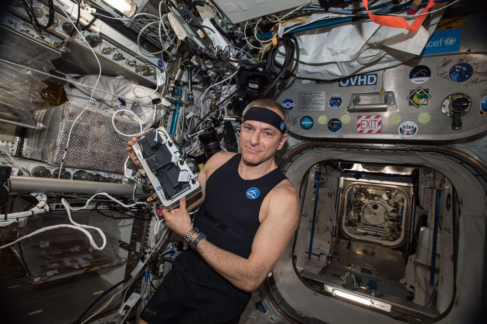Saint-Jacques wears a black sleeveless shirt and a black headband, both with a round blue CSA logo patch, and two watches on his left wrist. With both hands, he is holding a device the size of a large shoebox, with silver edges around black devices connected with several cords.