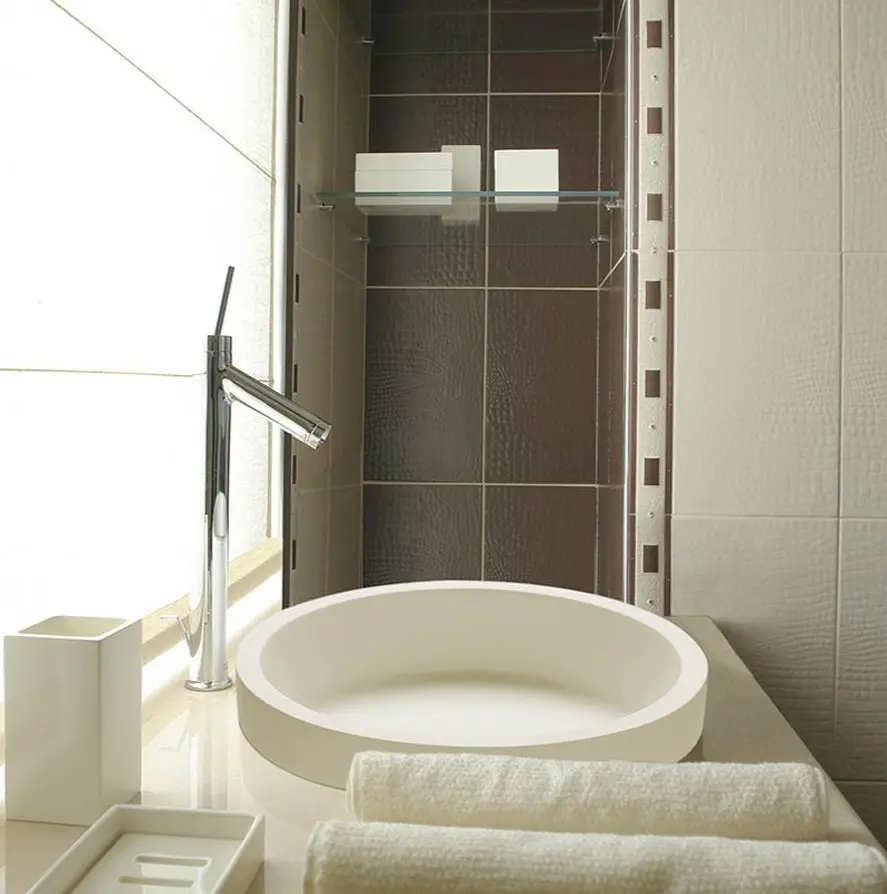 Circular semi-recessed bathroom sink in white; modern-looking stem-mounted faucet rises from the counter' towels in foreground, shower in background