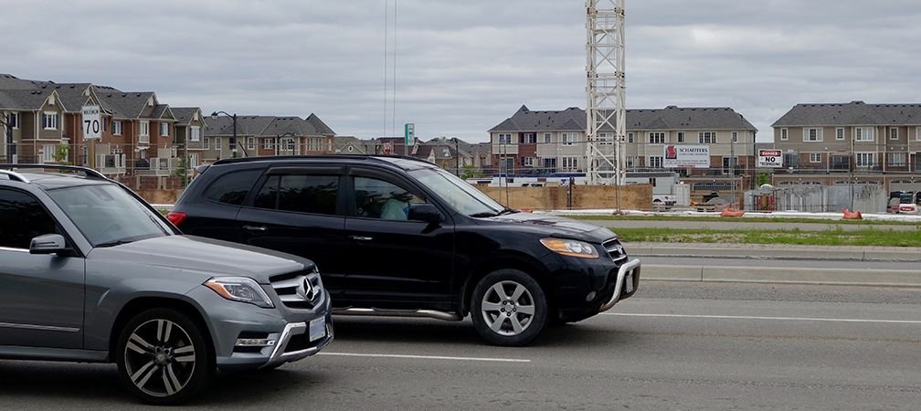 Two luxury vehicles drive by release emissions in front of a sprawling subdivision with thousands of residences packed closely together, human impacts at the cost of environmental degradation