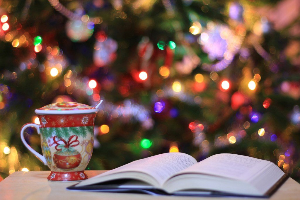 An open book is on a table next to a Christmas mug, in front of a Christmas tree lit by multicoloured Christmas lights. 