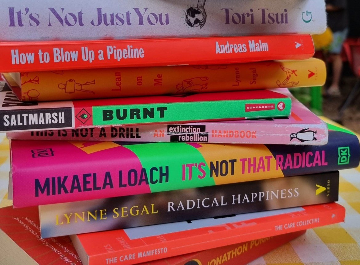 A tall pile of climate justice books on a chequered white and yellow table cloth. In the middle of the pile is Its Not That Radical by Mikaela Loach