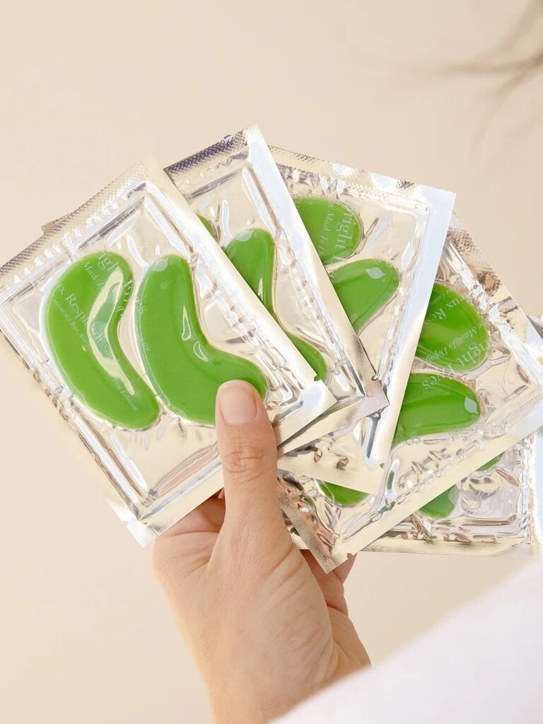 A hand holds four packages of green gel eye patches in silver foil wrappers, against a neutral background.