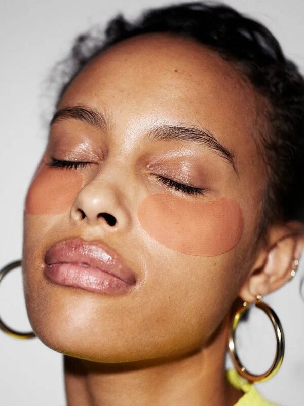 Person with closed eyes wearing under-eye patches, hoop earrings, and a light top, against a plain background.