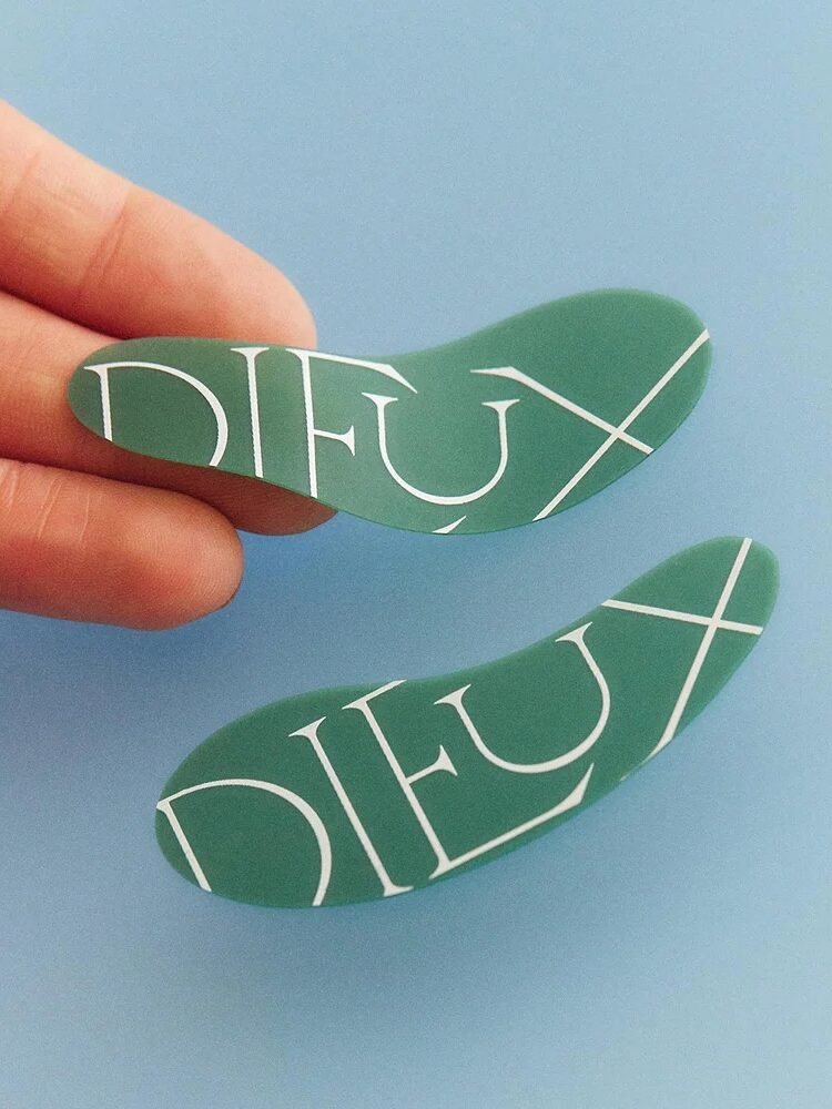 Hand holding two curved green eye patches with "DIEUX" printed on them against a blue background.