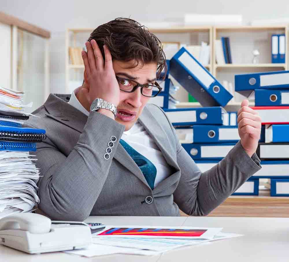 A stressed man in a suit sits at a cluttered desk with stacks of folders and paperwork marked "Certification." He holds a phone to his ear, gesturing frustration towards the papers. Shelves filled with more binders are in the background, hinting at endless tasks yet to be tackled.