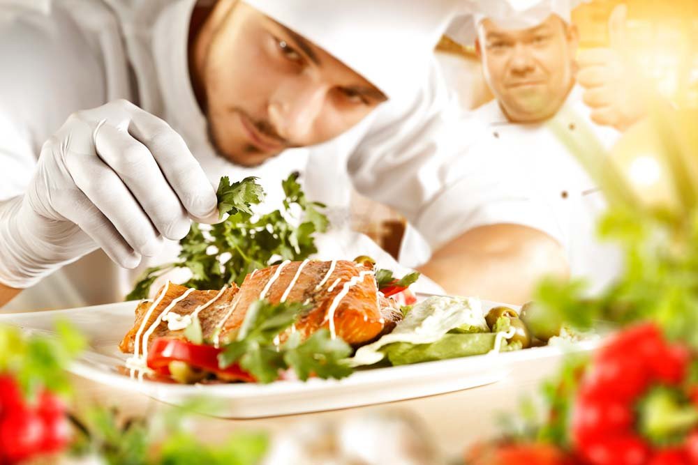 A chef in a white uniform and gloves carefully garnishes a salmon dish with herbs, embodying sustainable tourism practices. Another chef in the background gives a thumbs-up. The plate is surrounded by fresh vegetables, showcasing a culinary presentation that promotes GSTC principles.