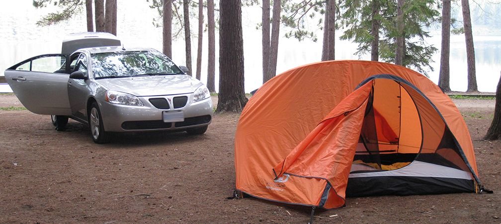 A car parked by a tent at a drive-in campsite in Algonquin Provincial Park nearby pine trees and a lake