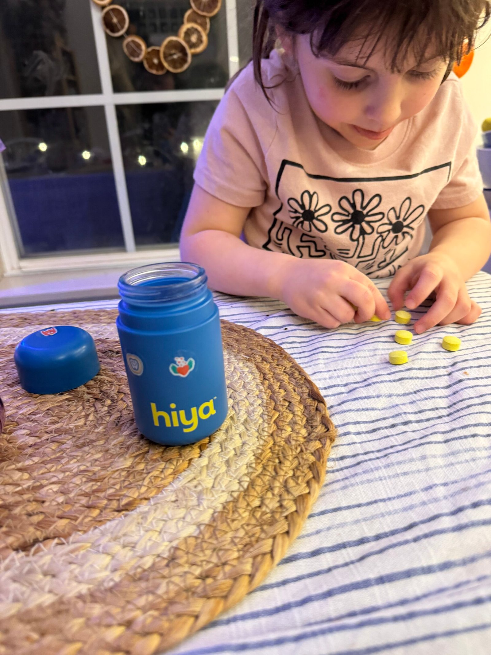 Child playing with yellow tablets next to a blue "hiya" bottle on a table with a striped cloth and woven mat.