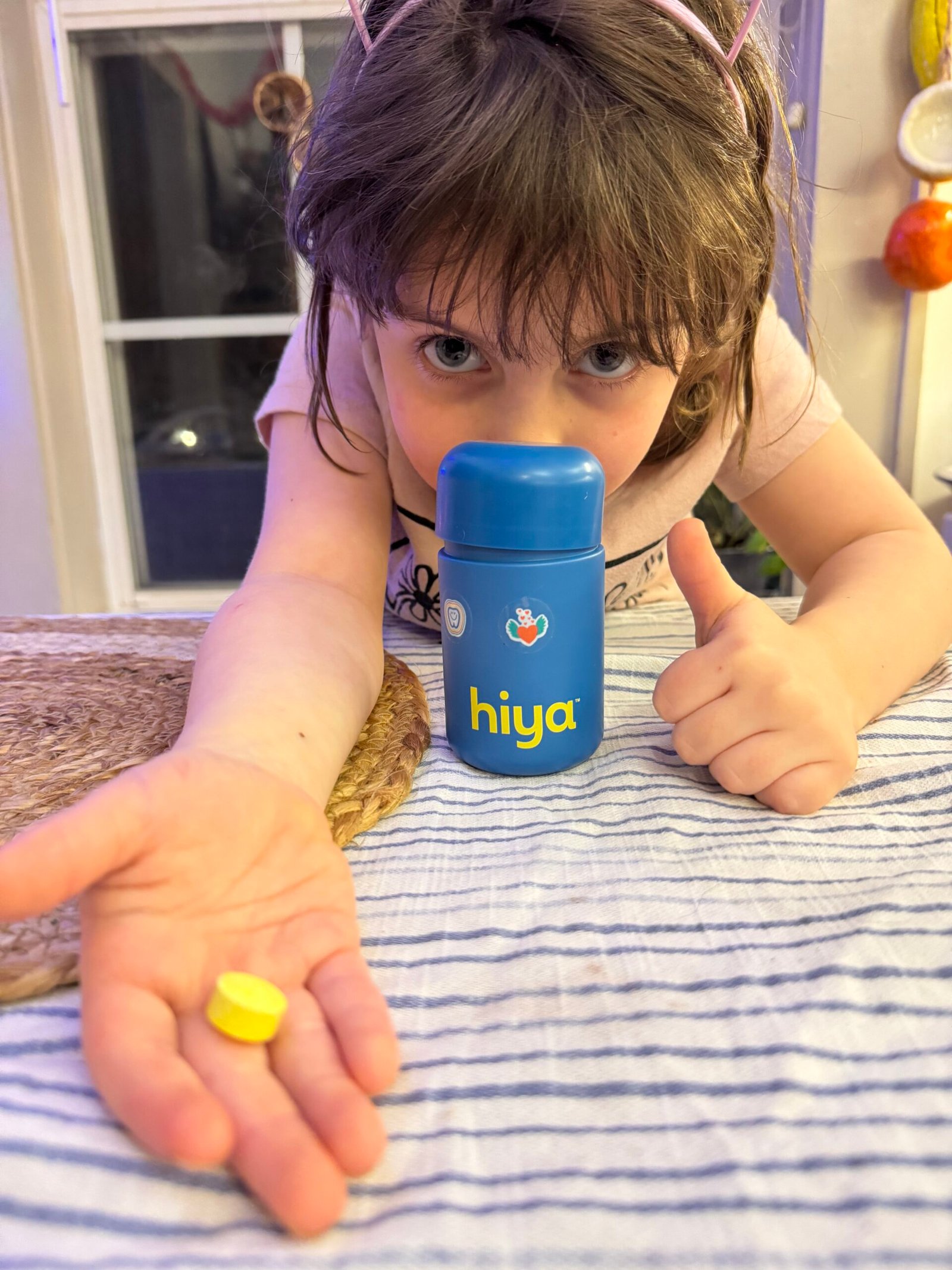 Child wearing cat ear headband giving a thumbs up, holding a yellow tablet, and pointing at a blue "hiya" bottle on a striped tablecloth.