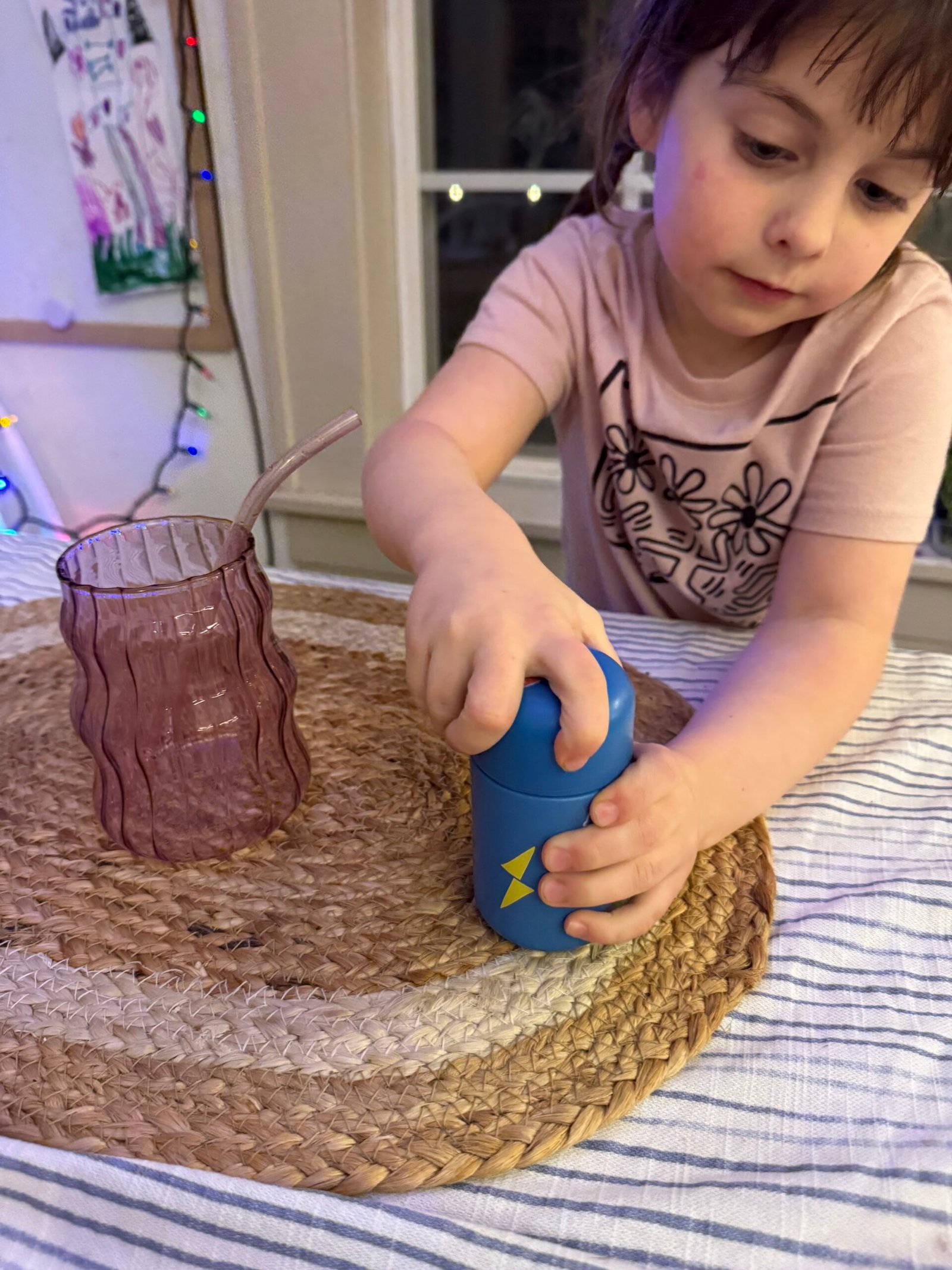 Child opening a blue can next to a purple glass on a woven mat.