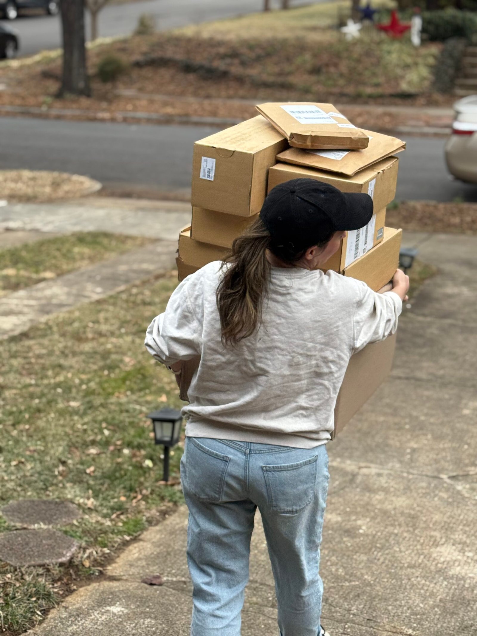 Person carrying a stack of cardboard boxes, wearing a black cap and gray sweatshirt, walking on a driveway.