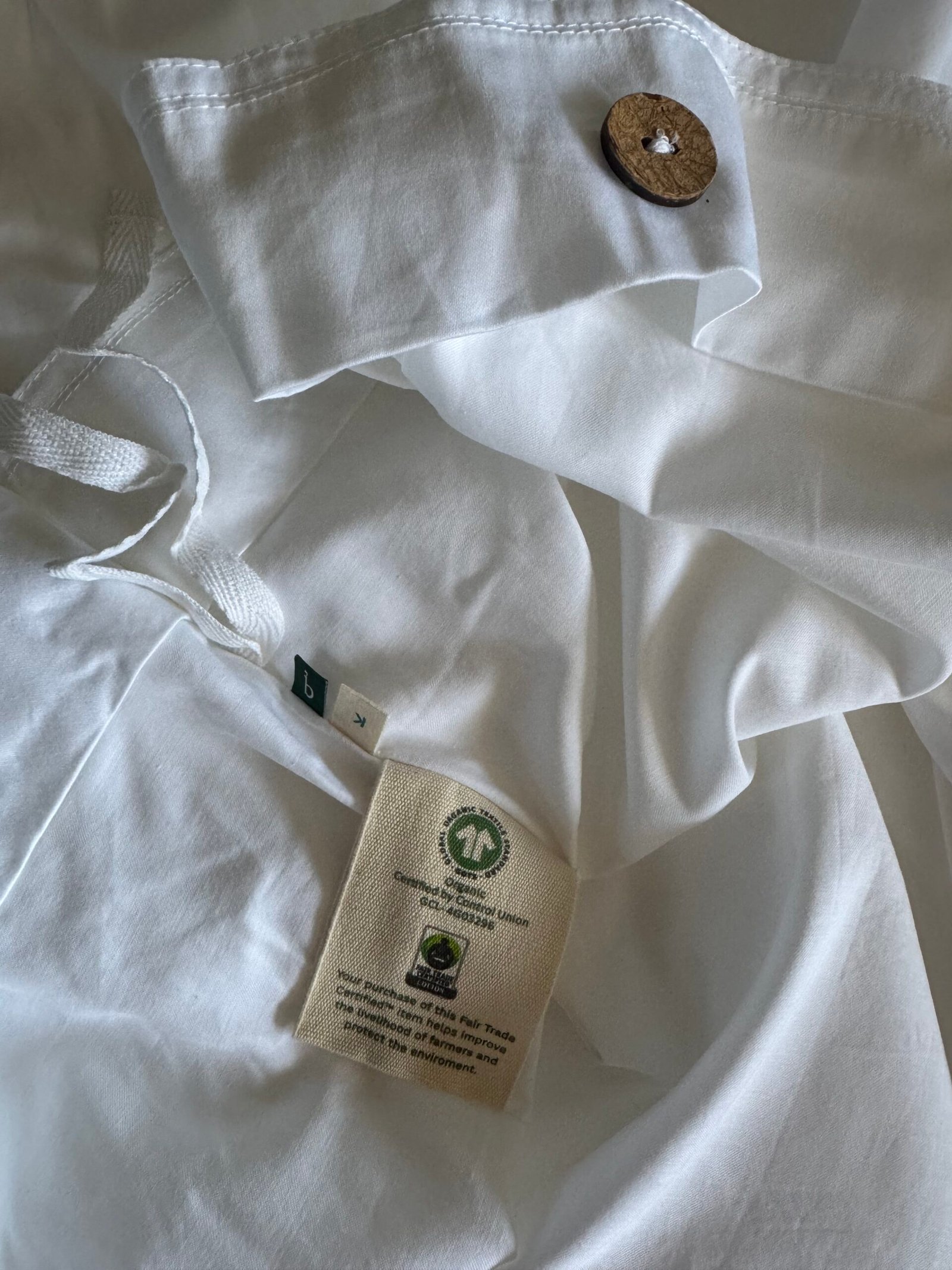 White fabric with a wooden button and multiple tags, including one indicating organic material and ethical certification logos.