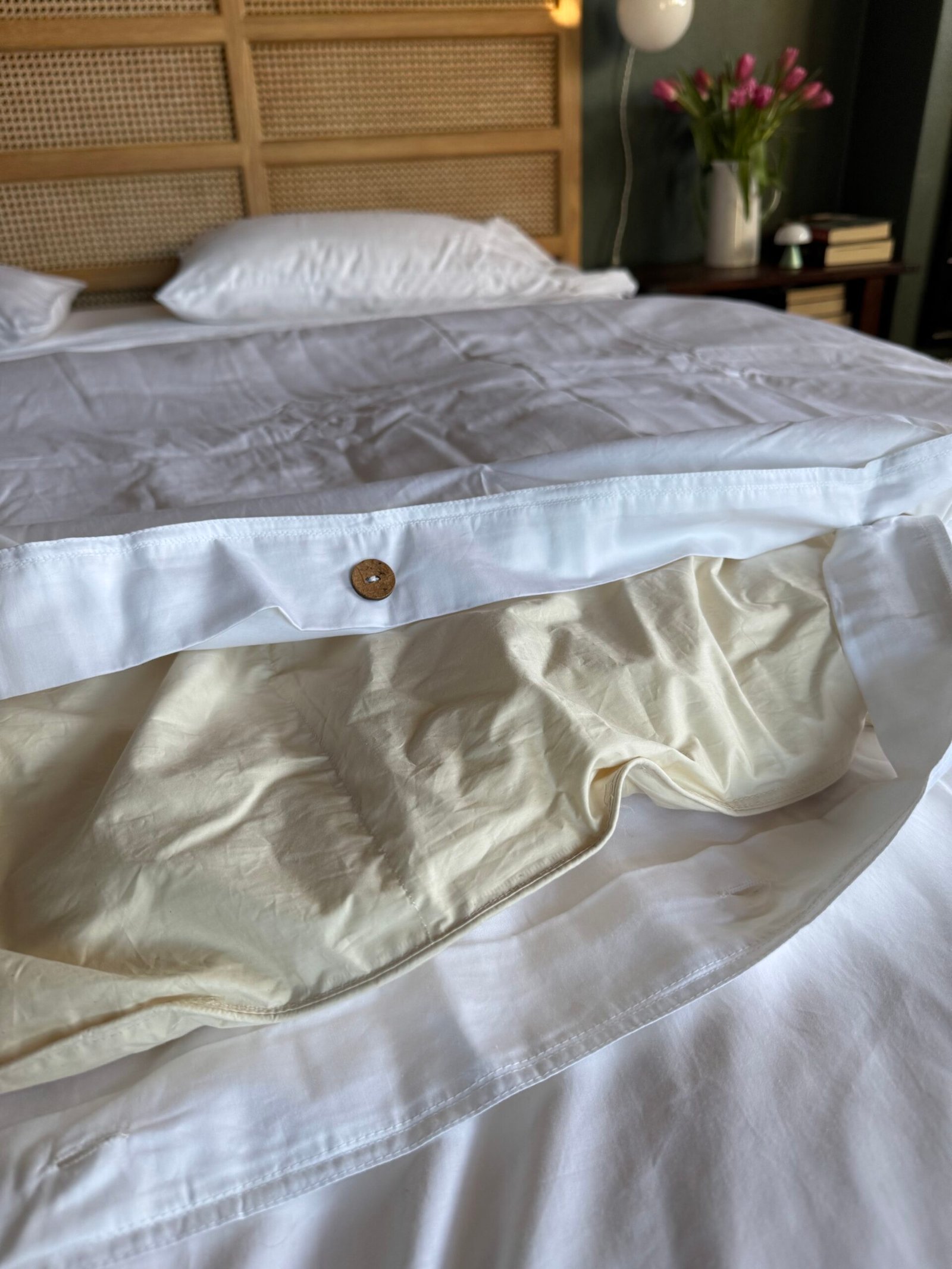 Unmade bed with disheveled white sheets and a cream comforter partially exposed. A button detail is visible on the duvet cover. Flowers and books are on a bedside table in the background.