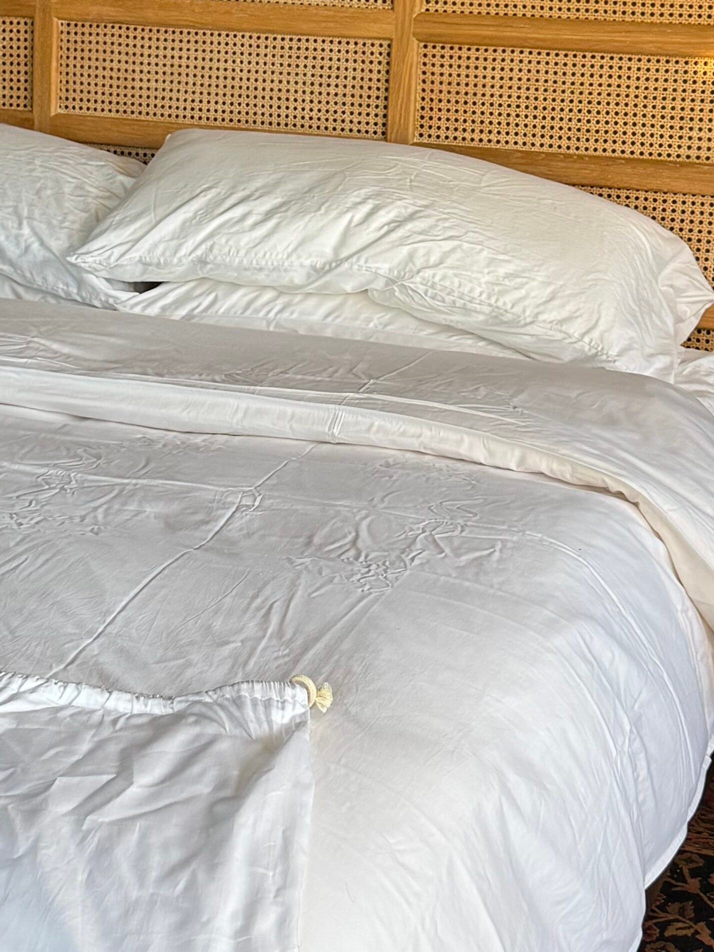 Bedroom with a double bed, white linens, and a rattan headboard. There are two bedside tables, flowers, a yellow vase, and a patterned rug.