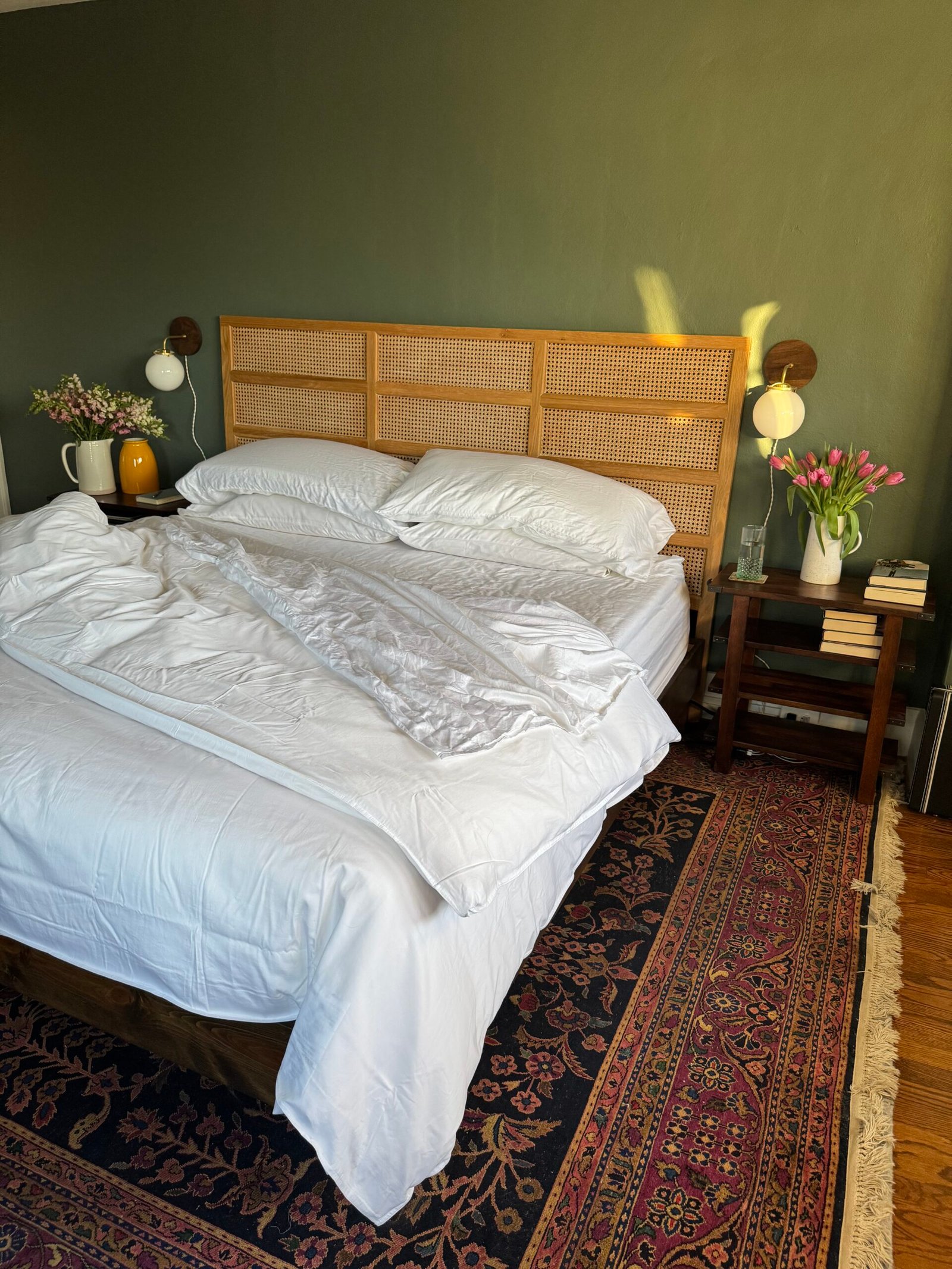 A neatly arranged bedroom with a rattan headboard, white bedding, and two wooden side tables holding lamps, flowers, and books. A patterned rug covers the hardwood floor.