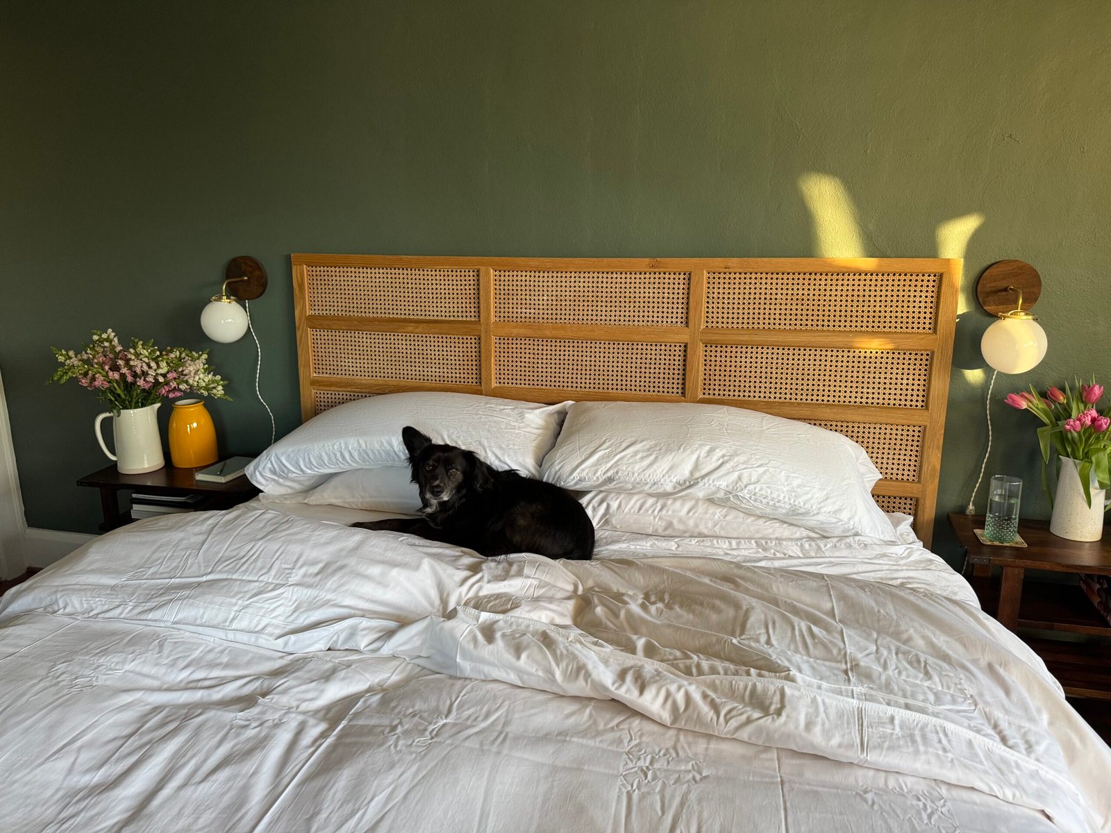 A black dog lies on a neatly made bed with white bedding. There are flowers and two lamps on the wooden side tables against a green wall.