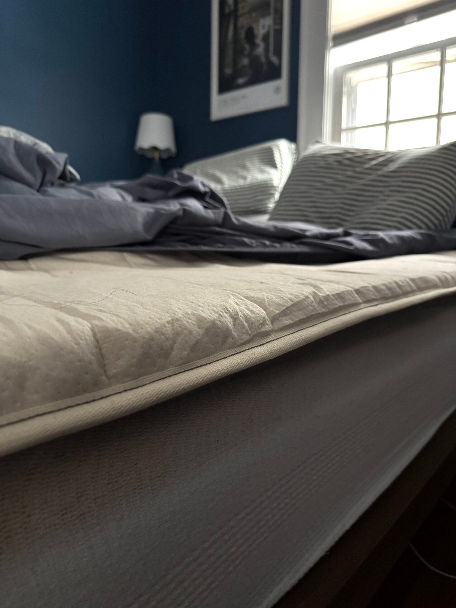 Unmade bed with rumpled sheets and pillows in a dimly lit room. Wall-mounted lamp and framed picture visible in background.