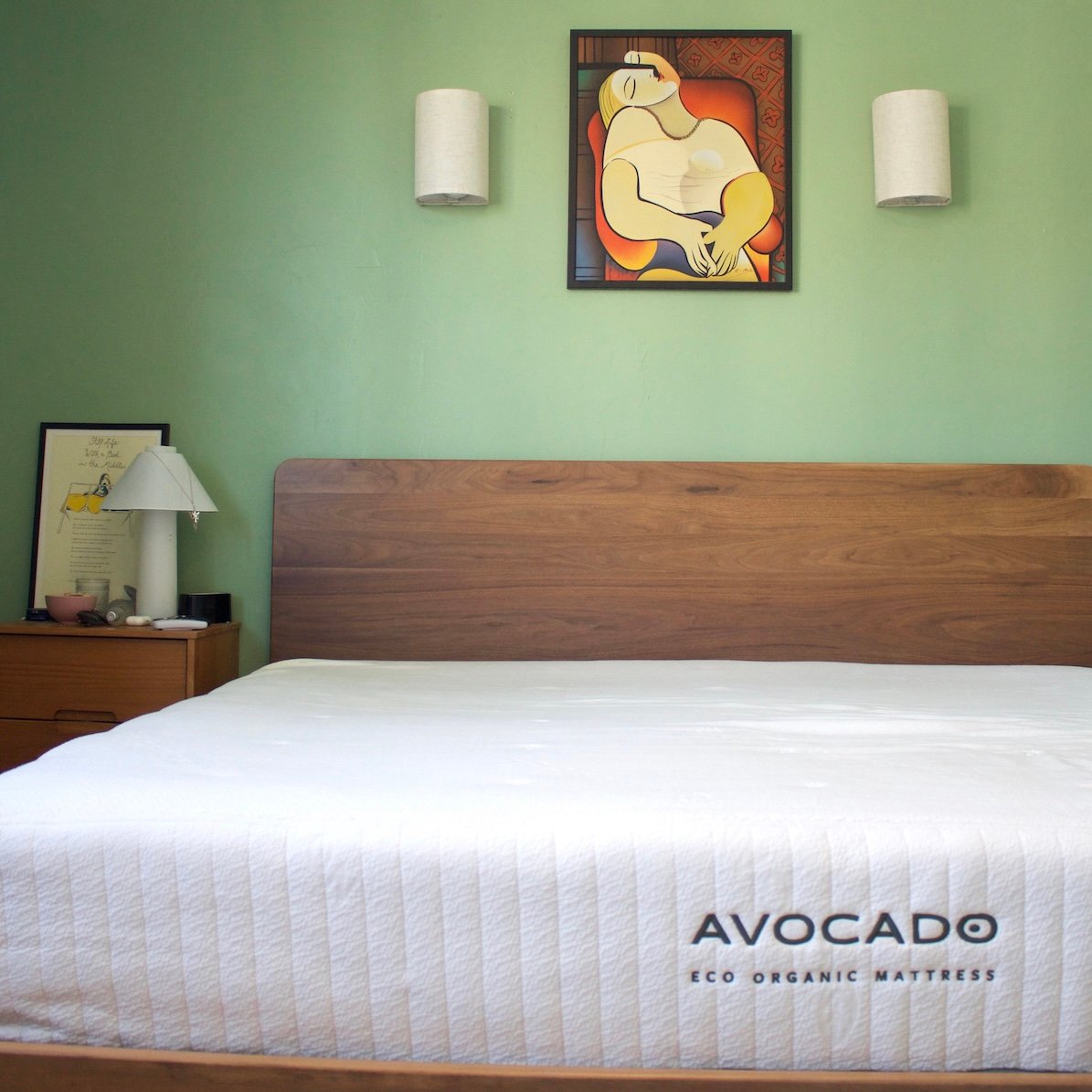 Bedroom with a wooden bed frame and white mattress labeled "Avocado Eco Organic Mattress." Light green wall with abstract art and two white wall lamps.