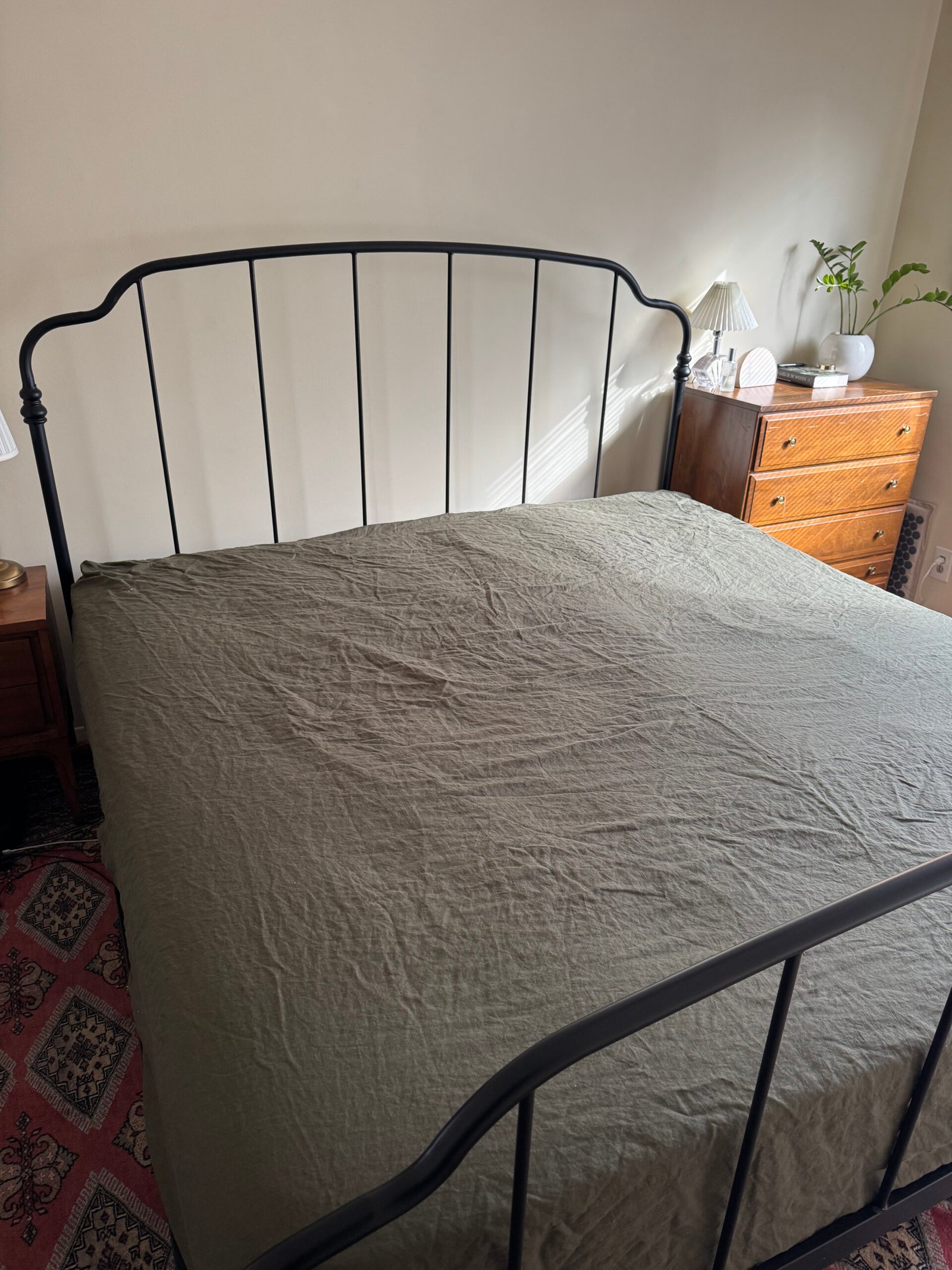 A bedroom with a metal bed frame, green bedspread, wooden dresser, lamp, and plant on top. A rug is on the floor.